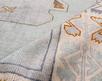 May include: A light blue area rug featuring a geometric pattern with gold, orange, and white accents. The rug is folded over to reveal a textured pile surface with a repeating border design and intricate linear motifs across the main field.