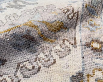 May include: A woven area rug featuring a cream-colored base with a repeating pattern in shades of muted blue, soft brown, and golden yellow. The texture is visible with a dense, knotted pile construction and a folded corner showing the backing.