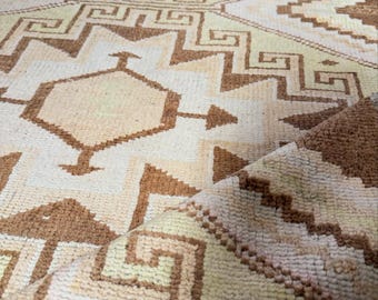 May include: A woven area rug featuring a repeating geometric pattern in shades of cream and light brown. The design includes interconnected diamonds, zig-zag lines, and Greek key motifs. The texture appears soft and tufted with a folded corner.
