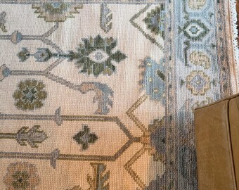 Peut inclure: Un tapis rectangulaire présentant un champ couleur crème avec un motif floral et géométrique répétitif dans des tons sourds de vert sauge, de bleu poussiéreux et de pêche doux. Le tapis possède une large bordure aux motifs complexes.