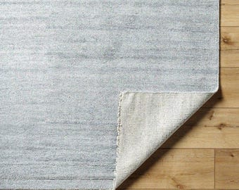 Puede incluir: Una alfombra rectangular azul claro con una superficie texturizada. La esquina de la alfombra está doblada, revelando el reverso blanco. La alfombra está sobre un suelo de madera.