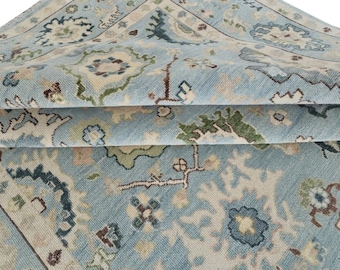 May include: A light blue area rug featuring a repeating floral and vine pattern in shades of cream, dark blue, green, and brown. The rug is folded over to reveal the textured weave and the intricate, symmetrical design across the surface.