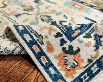 Peut inclure: Un tapis rectangulaire à fond crème orné d'un motif floral et géométrique multicolore dans des tons bleu foncé, orange, bleu clair et vert olive. Le tapis présente une texture épaisse et touffetée avec une bordure à franges sur le coin.