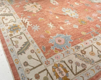 May include: A rectangular area rug featuring a terracotta orange field with a scattered pattern of stylized floral and geometric motifs in shades of light blue, cream, and sage green, framed by a wide cream border with intricate botanical designs.