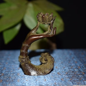 Könnte beinhalten: Eine bronzefarbene Skulptur einer Hand, die eine Lotusblume hält. Die Hand und die Blume sind detailliert, mit einem gebogenen Stiel und einem dekorativen Sockel. Der Hintergrund ist dunkel, mit grünen Blättern.