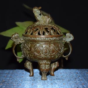 May include: A bronze-colored incense burner with intricate floral and scrollwork designs. The burner has a domed lid with openwork and a small animal figure on top. It features handles and rests on three feet.
