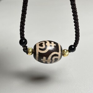 May include: A dark brown necklace featuring a cylindrical dzi bead with white and brown patterns. The bead is flanked by gold-colored beads and black beads, strung on a dark brown cord. The necklace is displayed against a neutral background.