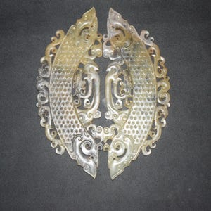 May include: Two symmetrical, light-colored, translucent stone carvings featuring intricate, openwork scroll patterns and a textured, scale-like design on the central curved sections. These decorative pieces exhibit varied shades of pale green, beige, and grey.