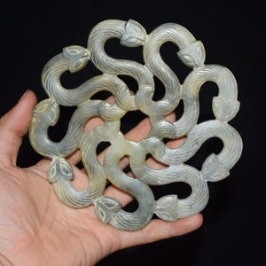 May include: A circular, pale green carved stone disc featuring an intricate, repeating pattern of intertwined, serpentine forms with pointed, leaf-like ends. The surface of the stone displays subtle, natural veining and a smooth, polished texture.