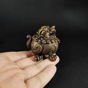 Peut inclure: Une figurine de créature mythique, possiblement un chien Foo ou lion gardien, de couleur bronze et aux détails complexes. La sculpture présente une crinière stylisée, une queue enroulée et des pattes détaillées. La figurine est tenue dans une main sur fond sombre.