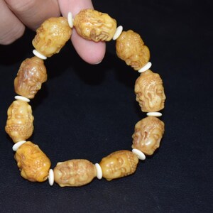 Peut inclure: Un bracelet composé de dix perles sculptées de couleur beige, chacune représentant un visage humain détaillé, alternant avec de petites perles d'espacement circulaires blanc cassé. Les perles sont enfilées sur un cordon élastique.