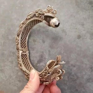 May include: A silver-colored hinged bracelet with a dragon design. The bracelet features intricate details, including a dragon head with a bell and floral patterns. The bracelet is open, revealing the inner lattice design.