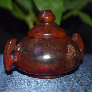 May include: A small, reddish-brown, lidded teapot-shaped container. The container has two handles and a rounded body. The lid has a small knob on top. The object is sitting on a blue patterned surface.