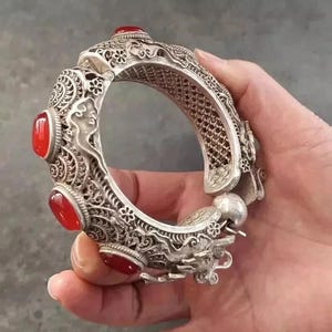 May include: Ornate silver bracelet with red gemstone accents. The bracelet features intricate floral and dragon designs, with a hinged clasp. The gemstones are oval-shaped and set into the silver. A detailed piece of jewelry.