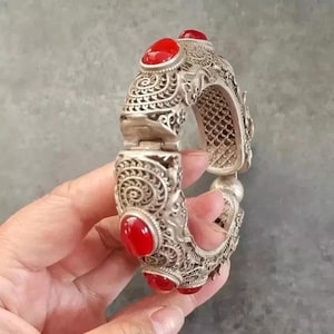 May include: Silver-toned hinged bracelet with intricate scrollwork and six oval, bright red gemstones. The bracelet is held open, showcasing the detailed design and the contrast between the metal and the red stones. A piece of jewelry.