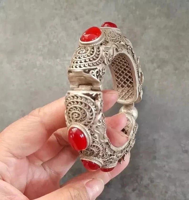 May include: Silver-toned hinged bracelet with intricate scrollwork and six oval, bright red gemstones. The bracelet is held open, showcasing the detailed design and the contrast between the metal and the red stones. A piece of jewelry.