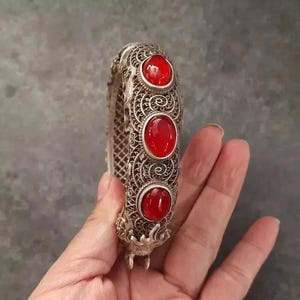 May include: Silver filigree bracelet with three large, oval, red gemstones. The bracelet has an ornate, openwork design and a clasp. The gemstones are set in decorative bezels. The bracelet is held against a neutral background.