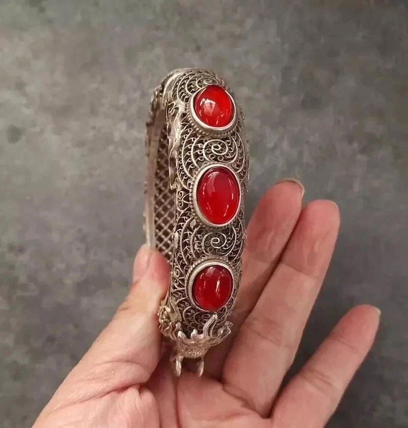 May include: Silver filigree bracelet with three large, oval, red gemstones. The bracelet has an ornate, openwork design and a clasp. The gemstones are set in decorative bezels. The bracelet is held against a neutral background.