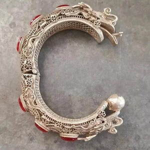 May include: Ornate silver-toned hinged bracelet with red gemstone accents. The bracelet features intricate filigree and dragon head details. The design includes floral and scroll patterns, creating a detailed and elegant accessory.