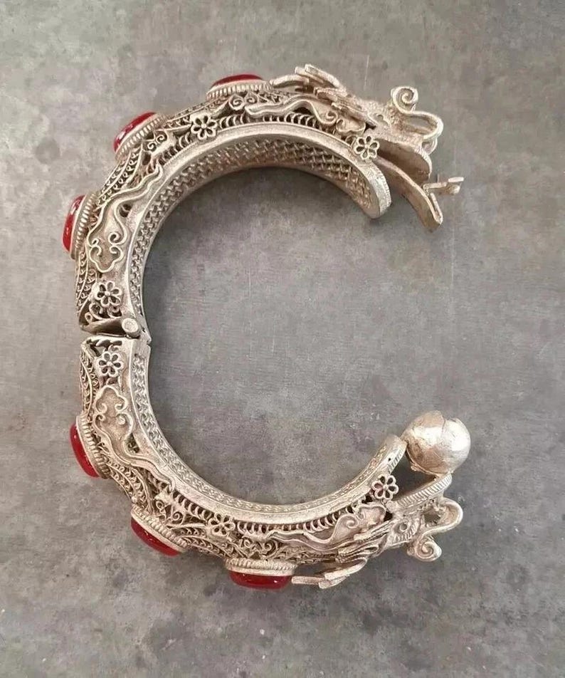 May include: Ornate silver-toned hinged bracelet with red gemstone accents. The bracelet features intricate filigree and dragon head details. The design includes floral and scroll patterns, creating a detailed and elegant accessory.