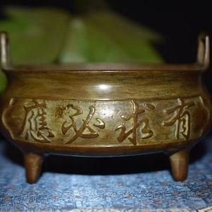 Könnte beinhalten: Ein bronzefarbener Räuchergefäß mit drei Beinen und zwei Griffen. Das Gefäß hat geprägte chinesische Schriftzeichen um den Körper. Der Hintergrund umfasst grünes Laub und eine blau gemusterte Oberfläche.