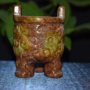 May include: A small, antique-style, brown and green carved vessel with a square shape and three legs. The vessel has two handles and is decorated with intricate patterns. The background is blurred, with green leaves and a blue surface.