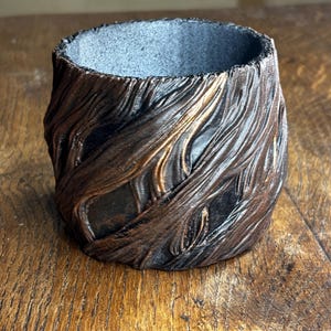 May include: A small, cylindrical container with a textured, wood-like appearance. The exterior features a dark brown and bronze colour scheme, with a rough, carved design. The interior is a contrasting grey colour. This decorative piece could be used as a small planter or a pen holder.