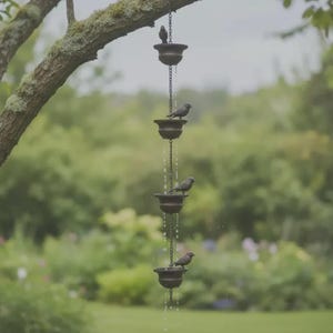 May include: A decorative rain chain with four bronze-colored bowls, each with a small bird perched on the edge. The chain hangs from a tree branch, with water flowing down the chain. The background is a blurred green garden.