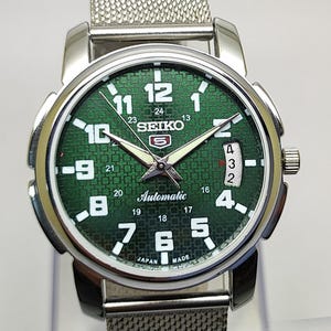 Könnte beinhalten: Eine silberfarbene Seiko Automatik-Armbanduhr mit grünem Zifferblatt. Die Uhr verfügt über ein Mesh-Armband, weiße Ziffern und eine Datumsanzeige. Das Zifferblatt enthält das Seiko-Logo und das Wort "Automatic".