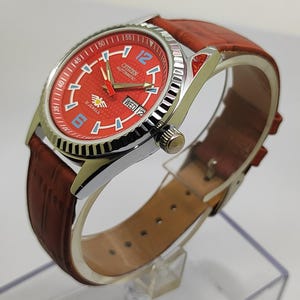 May include: A Citizen automatic wristwatch with a red dial and a brown leather strap. The watch has a silver bezel and features blue and white accents, including the number 12. The day and date are displayed at the 3 o'clock position.