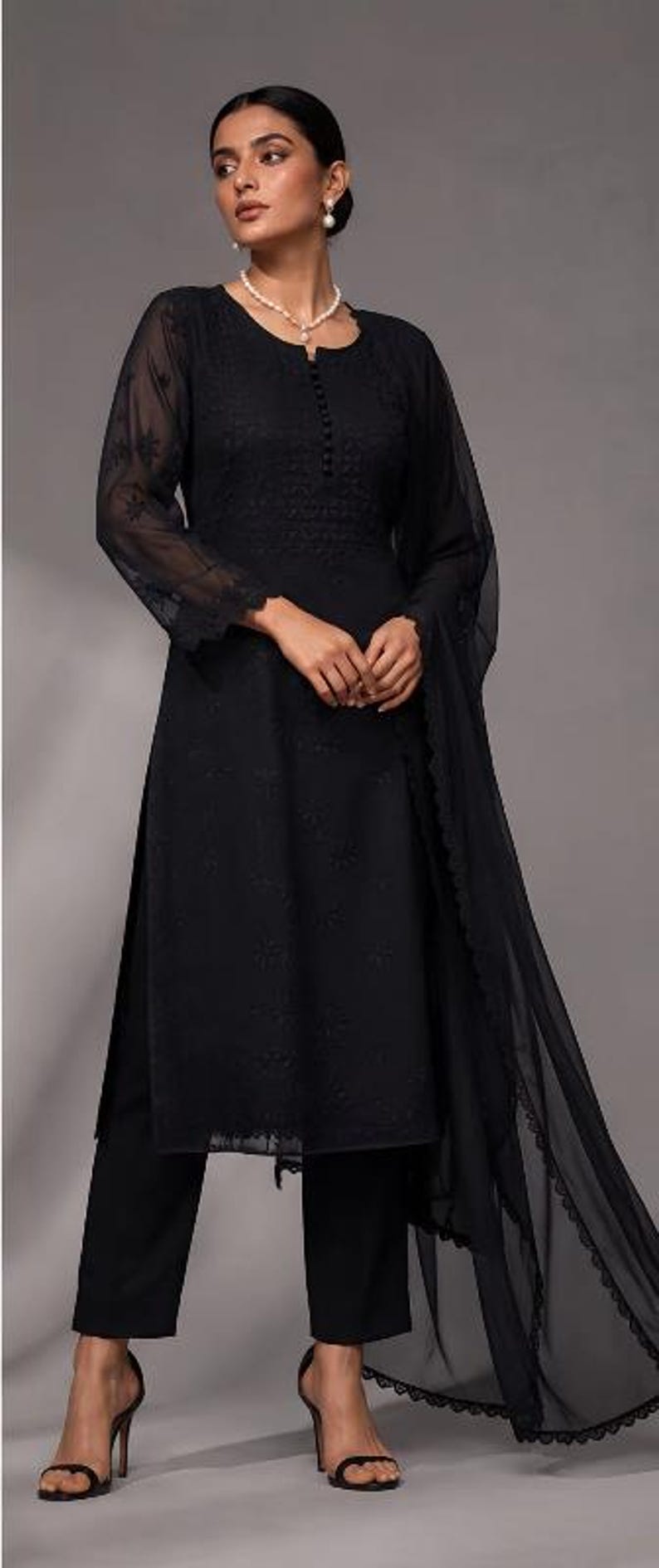 Puede incluir: Una t&uacute;nica negra de manga larga con bordados tonales intrincados, cuello redondo con tapeta de botones y pantalones rectos a juego. Un dupatta negro transparente con un borde de encaje festoneado completa el conjunto.