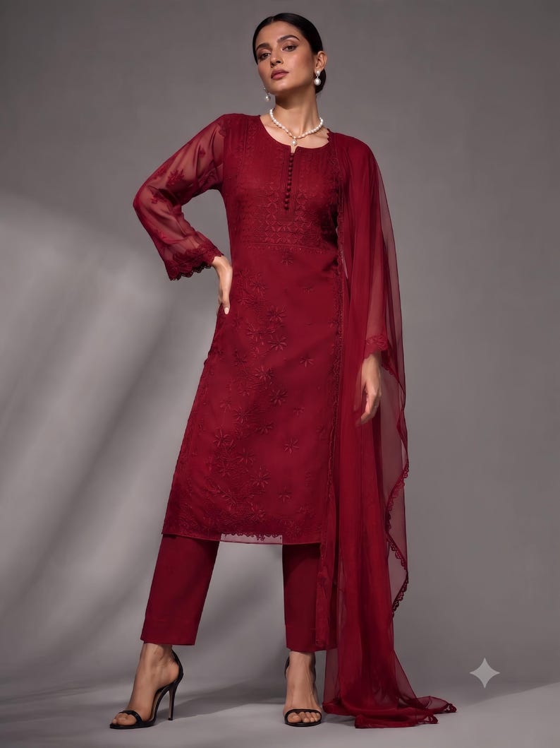 Puede incluir: Una t&uacute;nica de color granate de manga larga con intrincados bordados tonales y escote abotonado, combinada con pantalones rectos a juego y un dupatta ligero y transparente con un borde de encaje festoneado, usada con sandalias negras de tac&oacute;n.