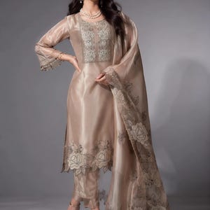 Puede incluir: Una túnica de organza beige con mangas tres cuartos, con intrincados bordados florales en el pecho y el dobladillo. El conjunto incluye pantalones rectos a juego y una dupatta transparente bordada drapeada sobre el hombro.