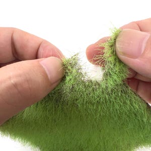 May include: Close-up of a vibrant green, textured material resembling grass or foliage. The material is held between two hands, revealing its detailed, fibrous structure. The color is a bright, natural green, suggesting its use in model making or crafting projects.