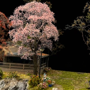 Op de afbeelding: Miniatuurscène van een huis met een veranda, een boom met roze bloesems en een stel. De scène bevat een lantaarnpaal, groen gras en kleurrijke bloemen. De achtergrond is zwart.