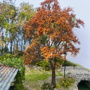 Puede incluir: Una escena de paisaje en miniatura con un árbol de follaje naranja-marrón, un puente de piedra y exuberante vegetación verde. Una farola negra se encuentra cerca del puente. La escena evoca el otoño.