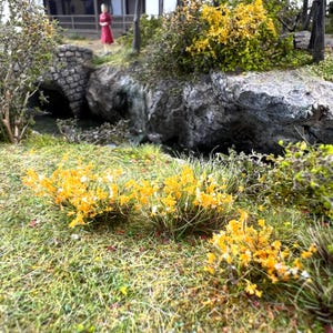 Può includere: Un diorama in miniatura che raffigura un ponte in pietra sopra un piccolo ruscello, circondato da erba verde e arbusti con fiori gialli. Una piccola figura in abito rosso si trova vicino al ponte, con alberi scuri e rocce sullo sfondo.