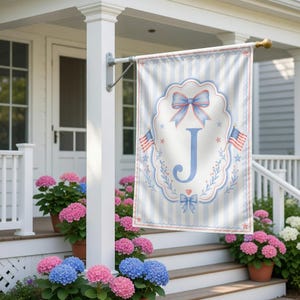 Puede incluir: Una bandera decorativa para exteriores con un fondo a rayas azul claro y blanco. La bandera presenta un diseño ovalado central con un lazo azul, la letra "J" y dos pequeñas banderas americanas. La bandera está colgada en un porche blanco.