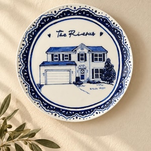 Puede incluir: Un plato decorativo redondo de cerámica blanca con una ilustración pintada a mano en azul de una casa de dos pisos con garaje. El texto "The Riveras" está escrito en letra cursiva azul sobre la casa, flanqueado por dos pequeños corazones azules.