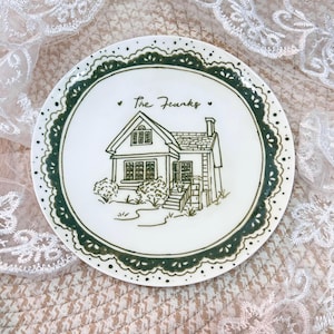May include: A white ceramic plate featuring a dark green hand-drawn illustration of a house with a chimney and steps. The text "The Franks" is written above the house. The plate has a decorative dark green scalloped lace-style border.