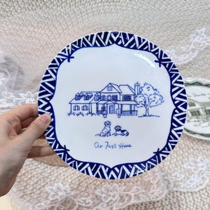 Puede incluir: Un plato redondo de cerámica blanca con un dibujo lineal azul de una casa con un árbol y dos perros. El texto "Our First Home" está impreso debajo de los perros. El plato tiene un patrón geométrico azul y blanco en el borde exterior.