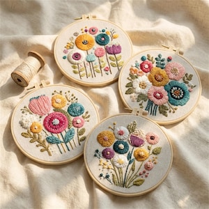 May include: Four circular wooden embroidery hoops containing fabric with colourful floral needlework designs in shades of yellow, pink, teal, and purple, accompanied by a small wooden spool of beige thread.