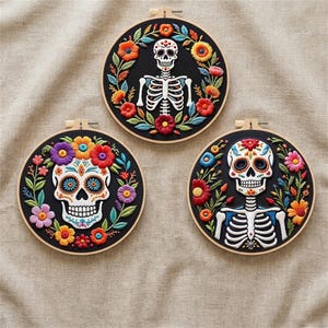 Könnte beinhalten: Drei runde Stickrahmen mit Totenkopf-Designs. Jeder hat einen schwarzen Hintergrund, farbenfrohe Blumenränder und einen detaillierten Schädel oder ein Skelett. Die Reifen sind auf einem neutralen Stoff platziert.
