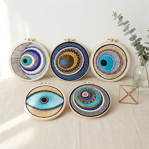 May include: A collection of five embroidered wall hangings featuring eye designs. Each piece is circular, set in a wooden hoop, and showcases intricate patterns in shades of blue, gold, and black. The designs vary, offering a unique aesthetic.