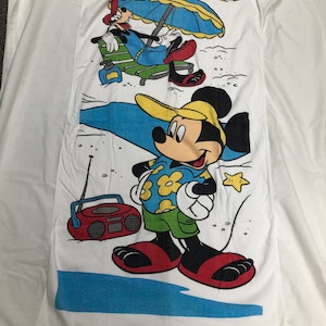 May include: A white beach towel with a cartoon image of Mickey Mouse and Goofy on a beach. Mickey Mouse is wearing a yellow hat, a blue and white Hawaiian shirt, green shorts, and red flip-flops. He is holding a white towel in his left hand. Goofy is wearing a red and yellow hat, a blue shirt, and blue pants. He is sitting in a green and white beach chair under a blue and yellow umbrella. There is a red and black boombox on the beach.