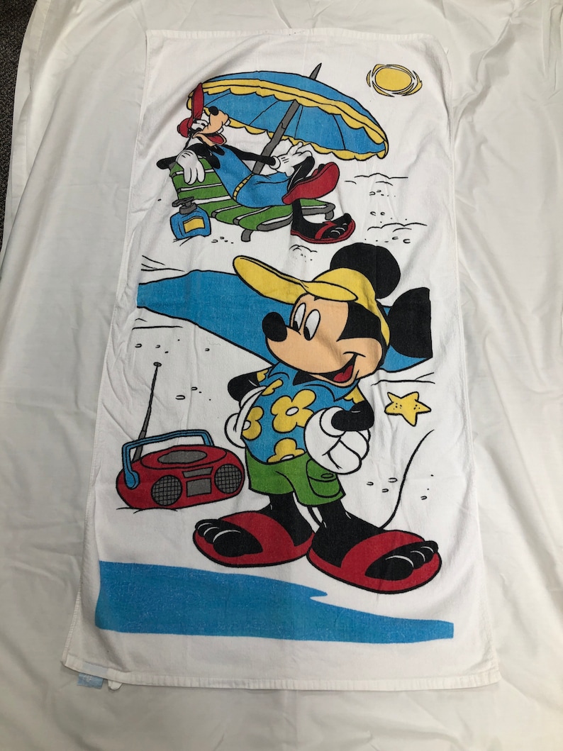 May include: A white beach towel with a cartoon image of Mickey Mouse and Goofy on a beach. Mickey Mouse is wearing a yellow hat, a blue and white Hawaiian shirt, green shorts, and red flip-flops. He is holding a white towel in his left hand. Goofy is wearing a red and yellow hat, a blue shirt, and blue pants. He is sitting in a green and white beach chair under a blue and yellow umbrella. There is a red and black boombox on the beach.