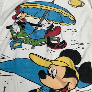 May include: A white beach towel with a cartoon image of Mickey Mouse and Goofy. Mickey Mouse is wearing a blue and yellow shirt with yellow flowers and a yellow hat. Goofy is wearing a blue and green shirt and red shoes. Goofy is sitting in a green and white lounge chair under a blue and yellow umbrella. The towel has a yellow sun and a starfish.