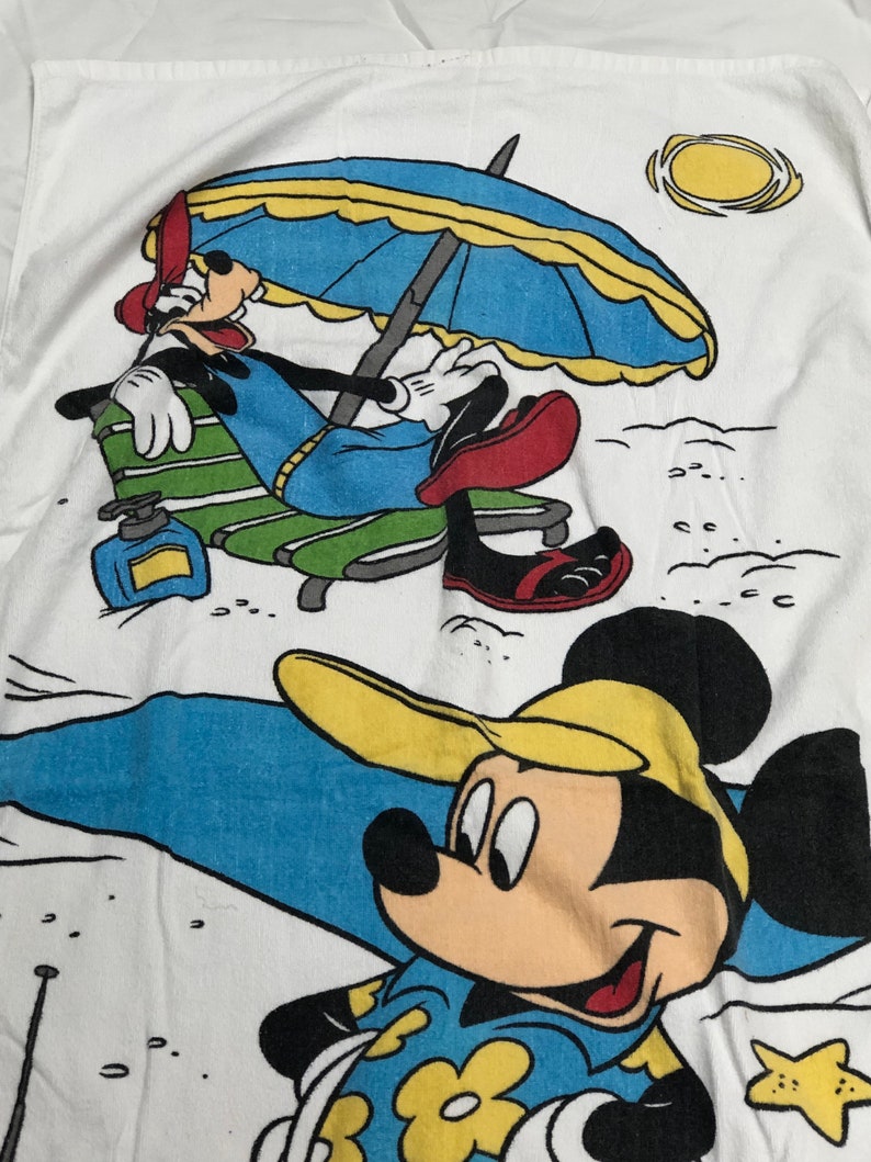 May include: A white beach towel with a cartoon image of Mickey Mouse and Goofy. Mickey Mouse is wearing a blue and yellow shirt with yellow flowers and a yellow hat. Goofy is wearing a blue and green shirt and red shoes. Goofy is sitting in a green and white lounge chair under a blue and yellow umbrella. The towel has a yellow sun and a starfish.