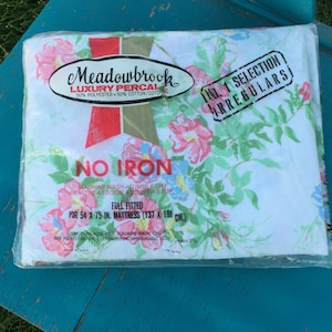 Puede incluir: Una sábana bajera blanca con estampado floral en tonos rosa, verde y azul. La sábana está etiquetada como "Meadowbrook Luxury Percale" y "No Iron". Es una sábana de tamaño completo, 54 x 75 pulgadas (137 x 190 cm).
