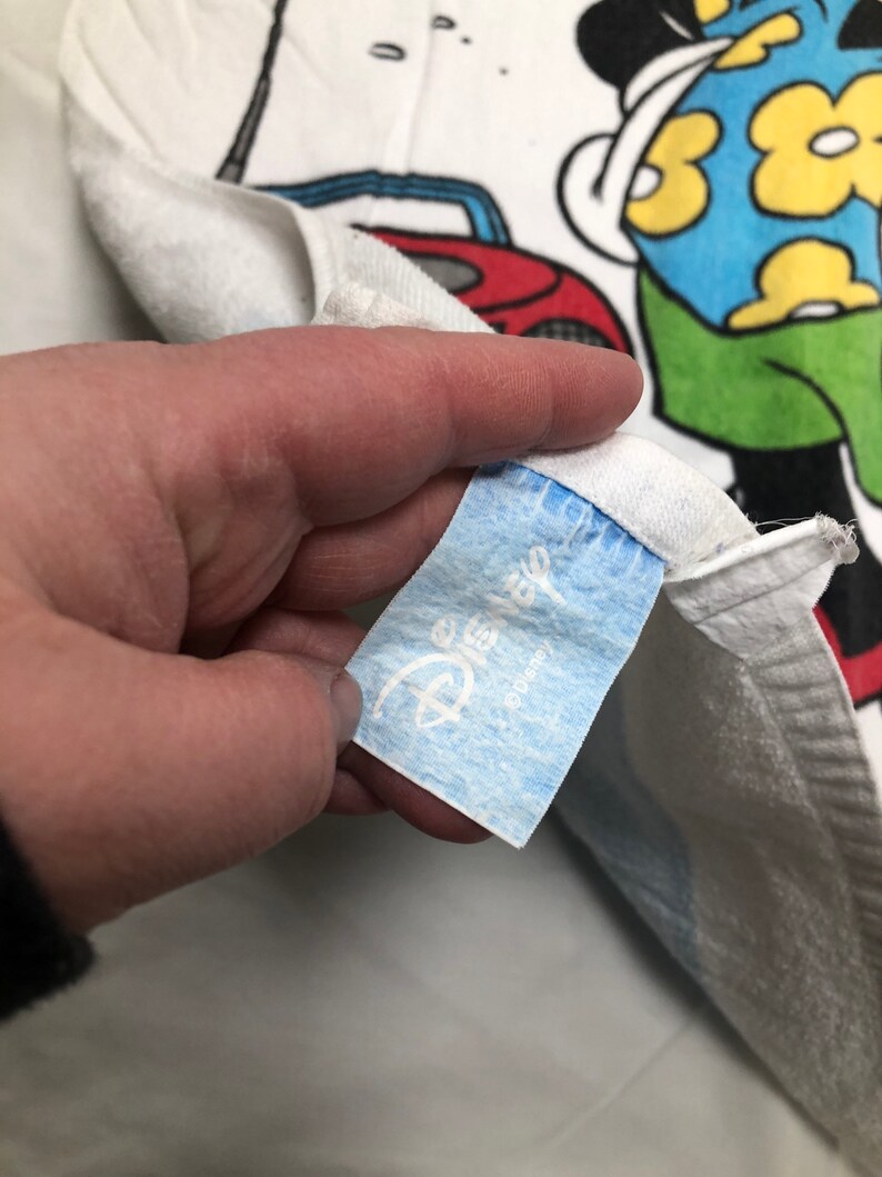 May include: White towel with a blue and yellow cartoon character wearing a blue shirt and green shorts. The character is holding a golf club and there is a red car in the background. The towel has a blue Disney label with the word "Disney" in white.